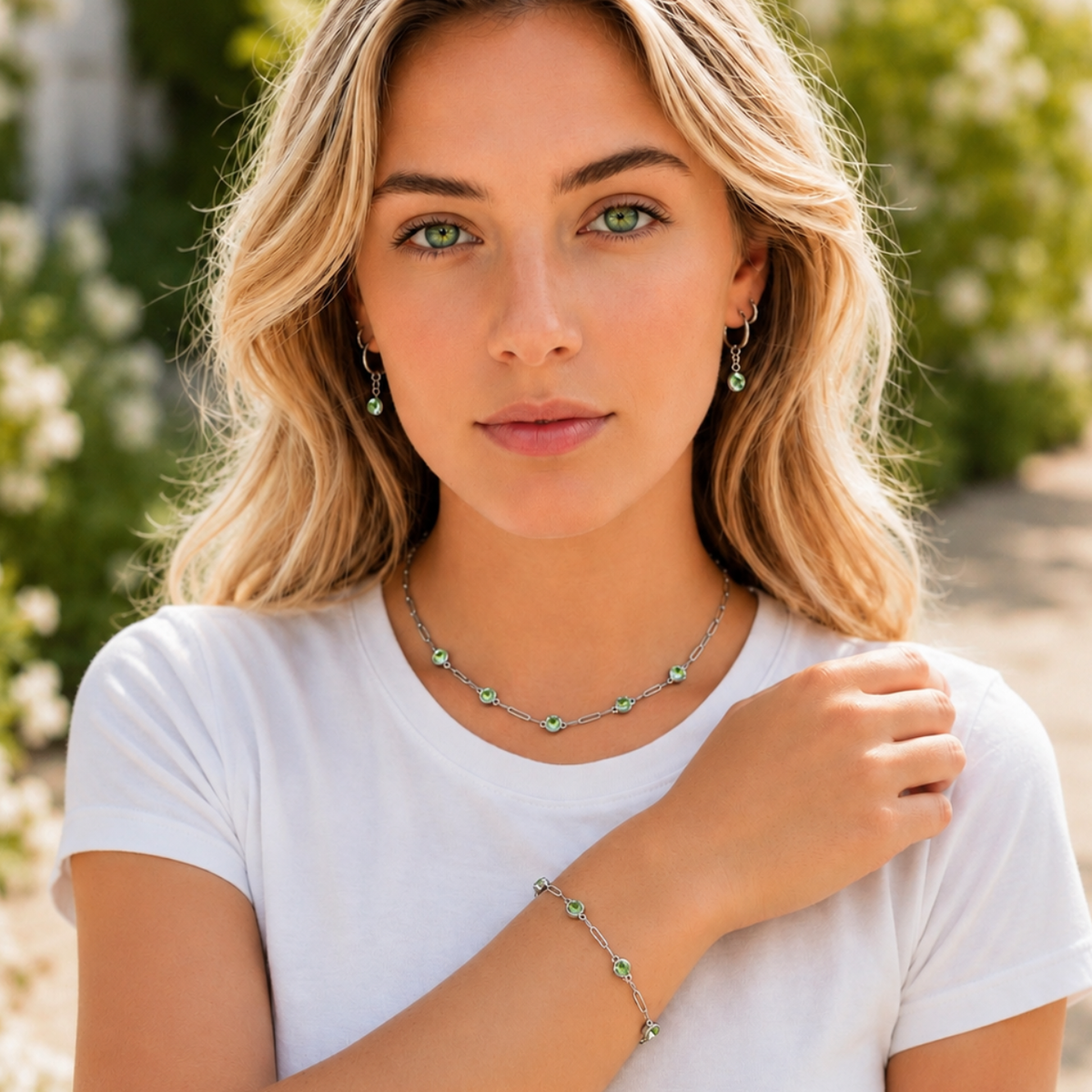 A blonde woman wearing a silver-toned necklace, matching bracelet, and drop earrings with small green stones, photographed outdoors with blurred greenery in the background.
