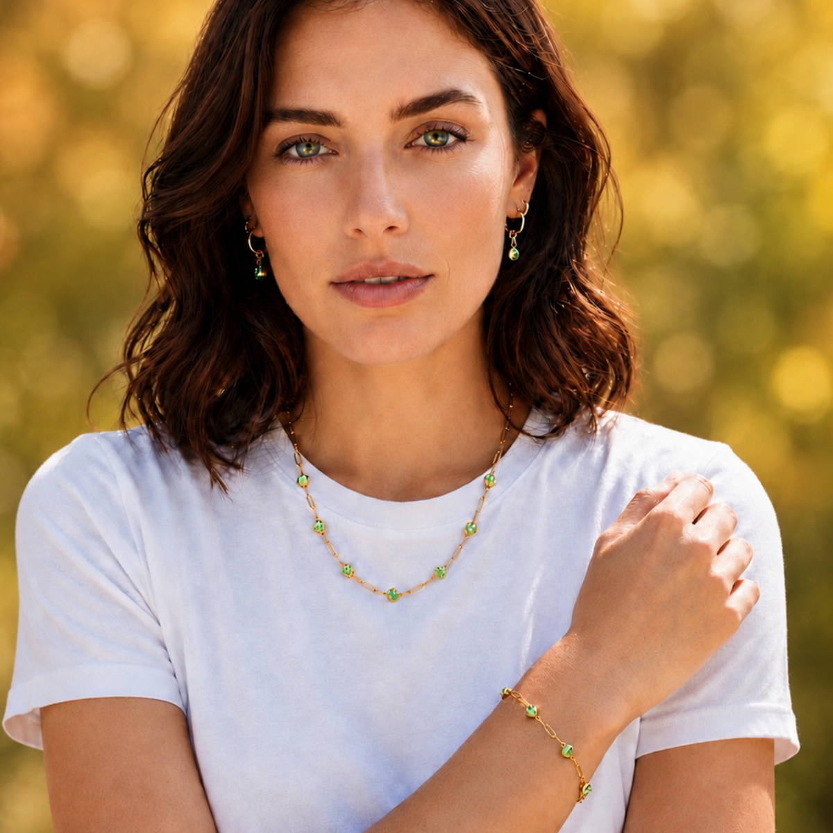 A woman wearing a delicate gold necklace and matching bracelet with small green gemstone accents, styled with matching drop earrings, set against a soft, blurred yellow-green bokeh background.
