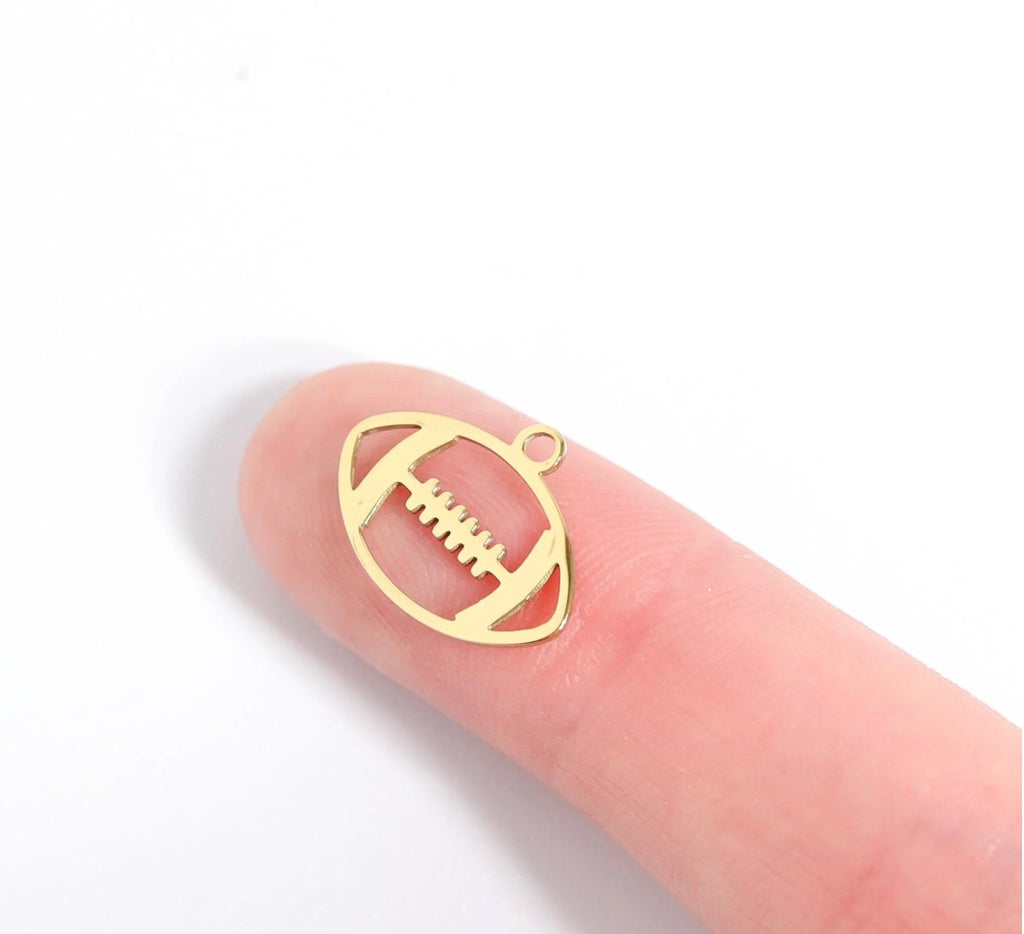 A small gold-colored football-shaped jewelry charm or pendant displayed on a person's fingertip against a plain white background.