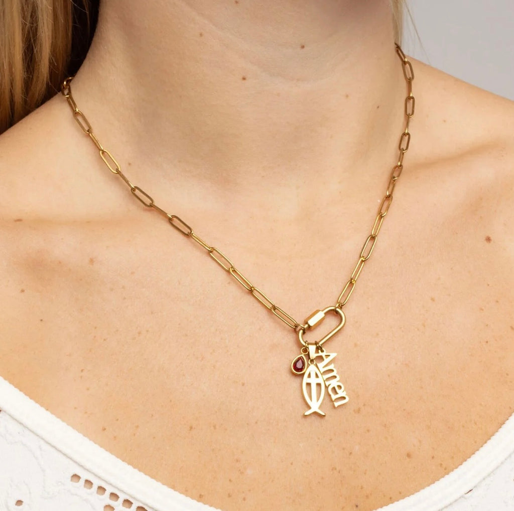 A gold paperclip chain necklace with a small dangling fish charm and a tiny red heart accent, shown worn on a model's neck.