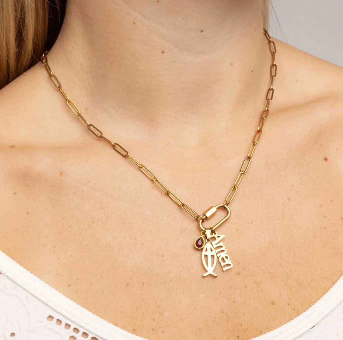 A gold paperclip chain necklace with a small dangling fish charm and a tiny red heart accent, shown worn on a model's neck.