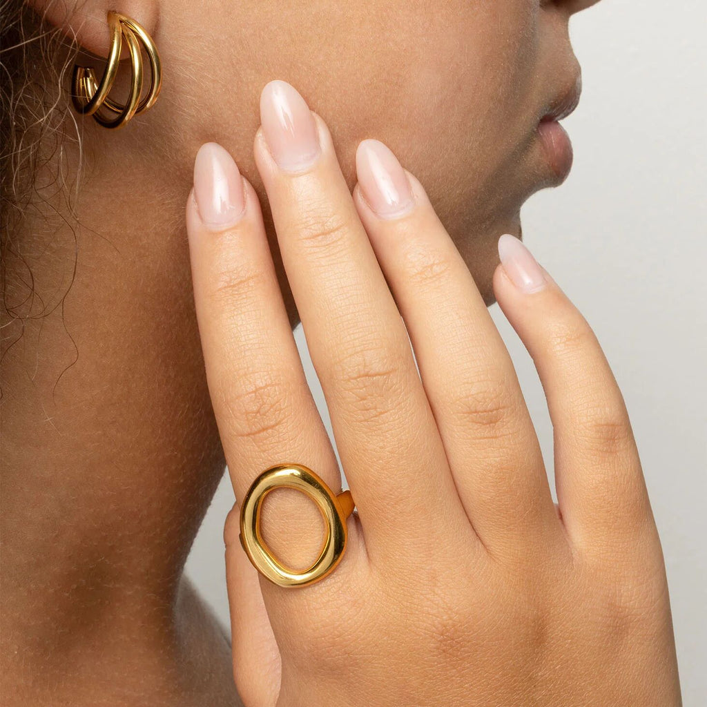 Close-up of a person wearing gold hoop earrings and a gold ring on a neutral background