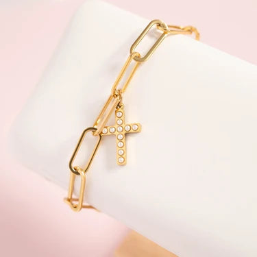 A gold chain bracelet with a dangling cross charm, displayed on a white jewelry stand against a soft pink background.