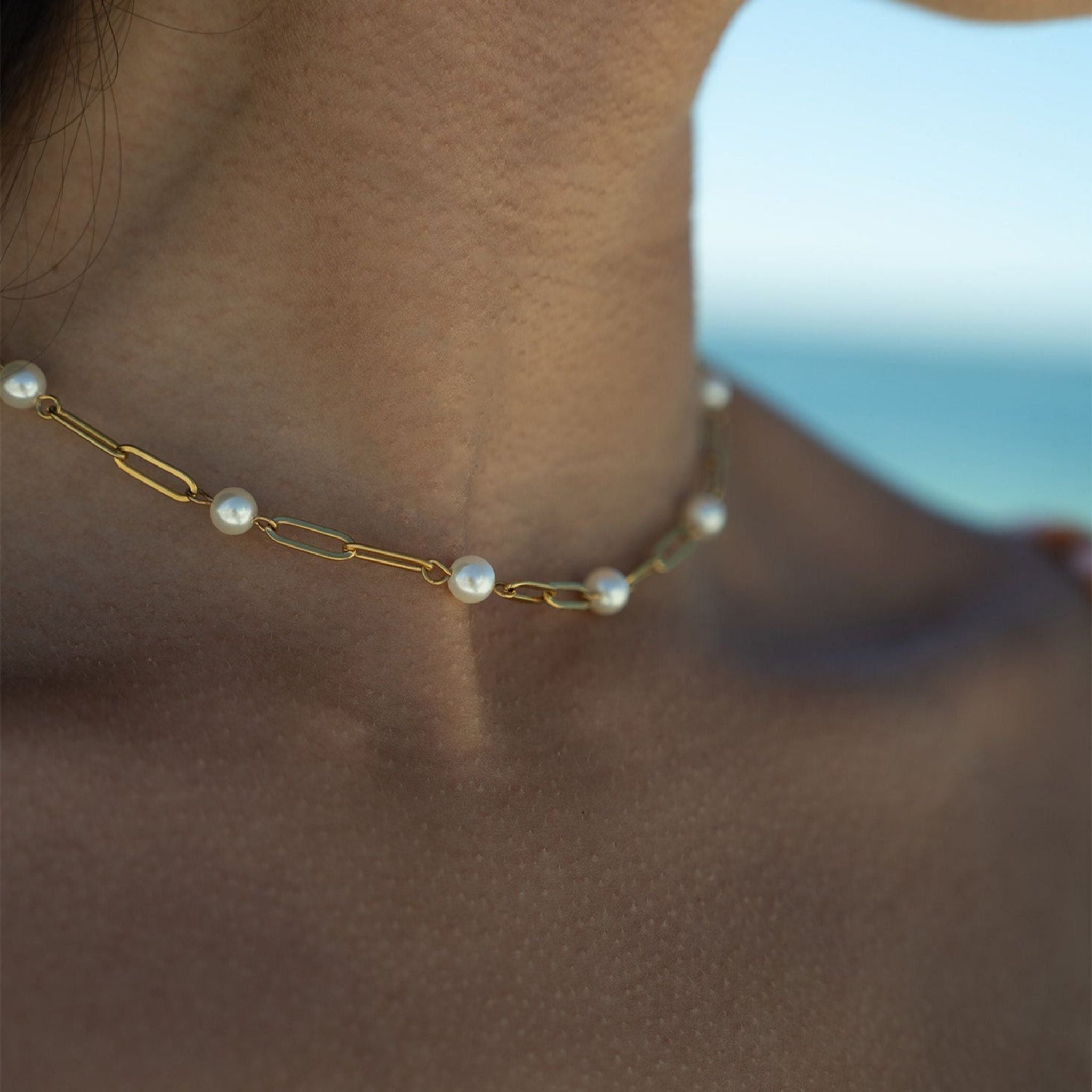 A close-up of a delicate gold chain necklace adorned with small white pearls, worn on a model's neck with a blurred blue ocean background.