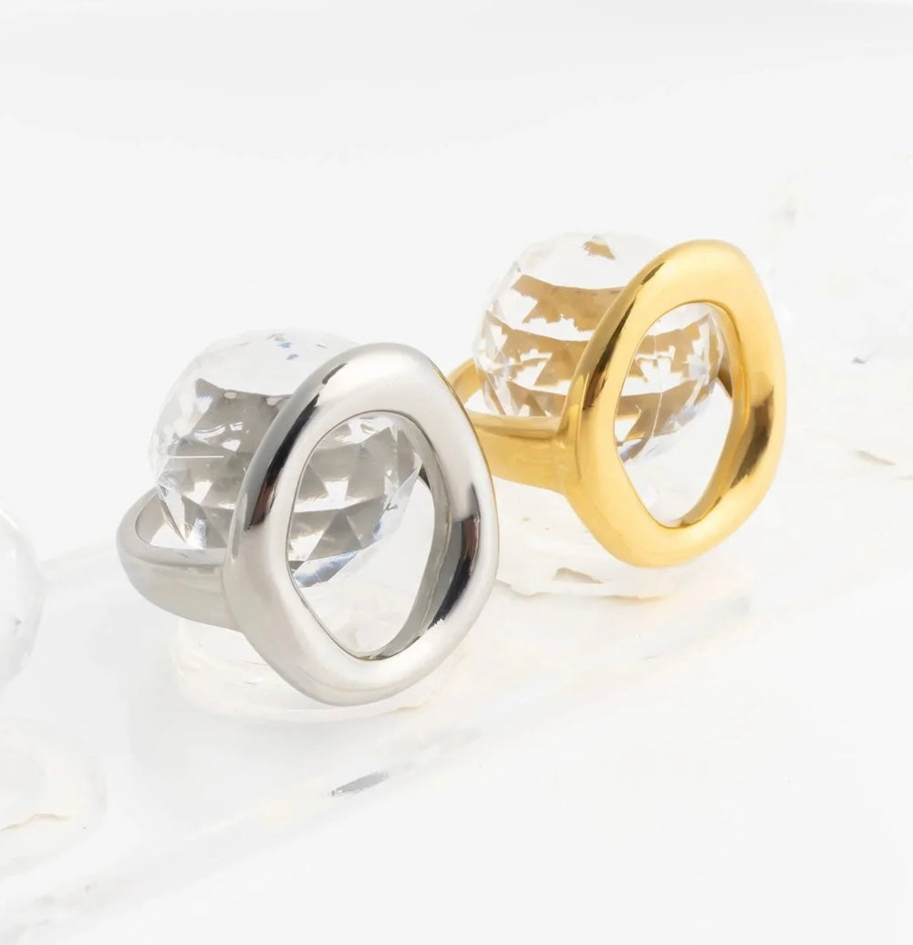 Two oval ring-shaped earrings with faceted clear crystal centers, one in polished silver-tone metal and the other in polished gold-tone metal, displayed on a white reflective surface.