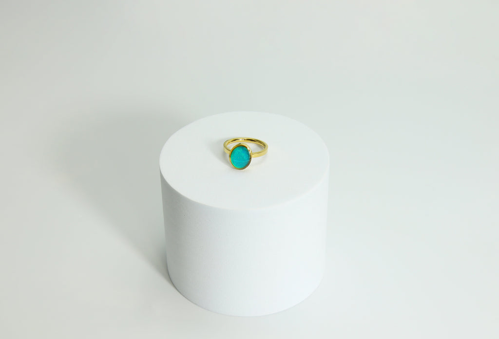 Gold mood  ring with a turquoise gemstone on a white cylindrical stand against a light gray background