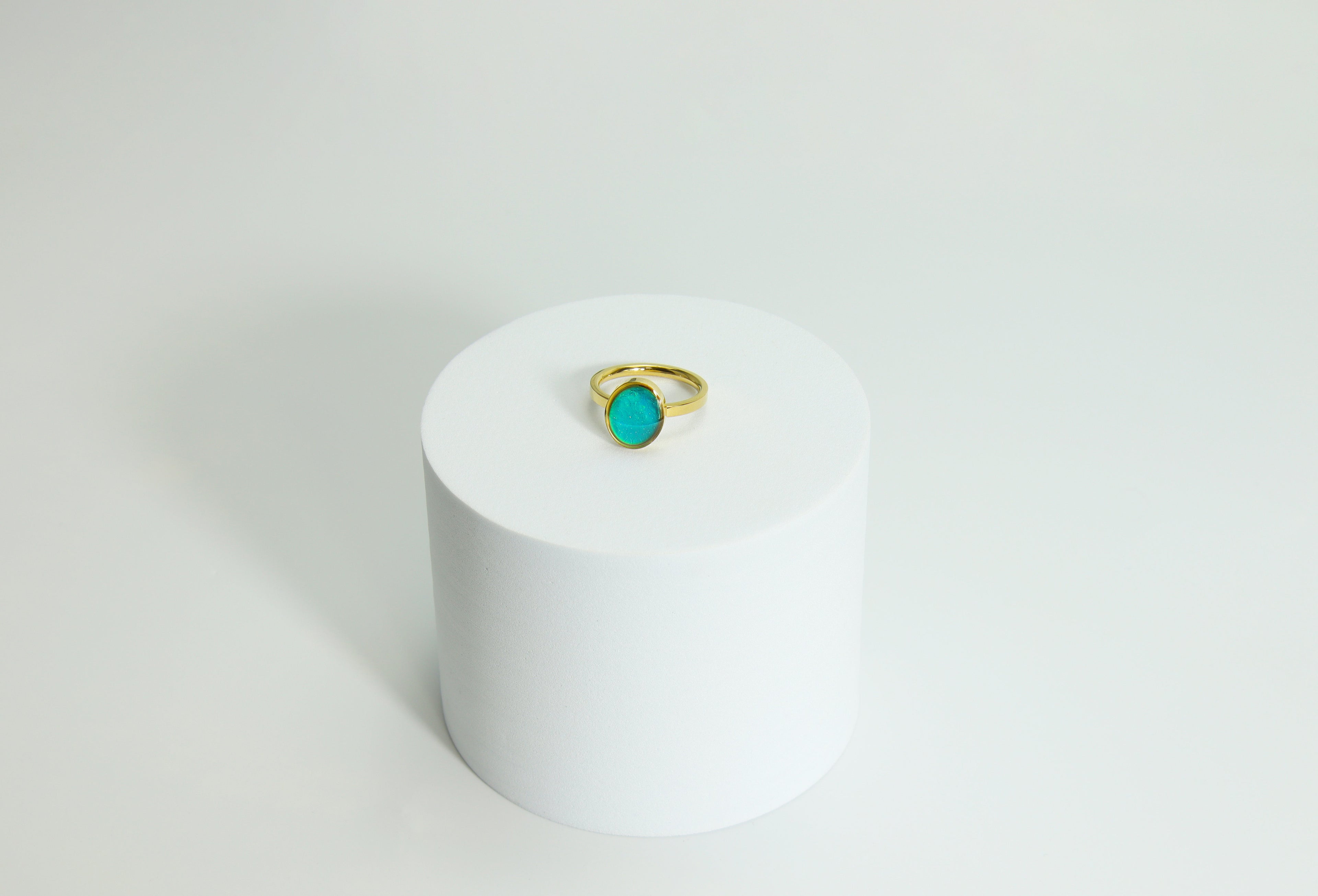 Gold mood  ring with a turquoise gemstone on a white cylindrical stand against a light gray background