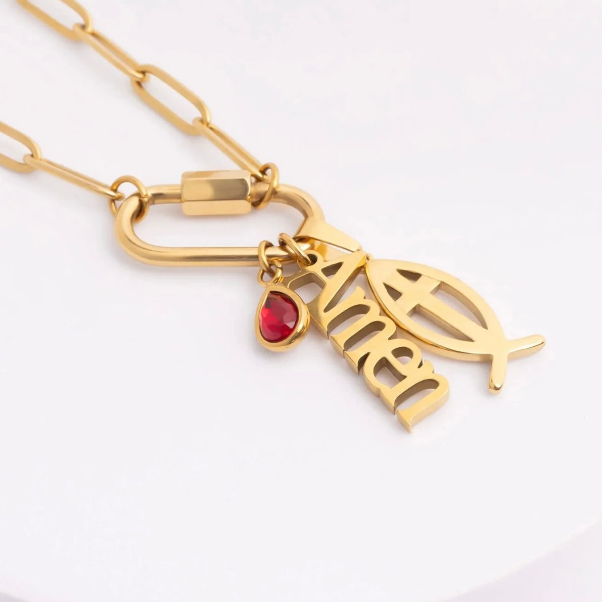 Gold-tone chain necklace with a clasp and three charms: a fish symbol, a stylized nameplate reading 'Amen', and a small red heart-shaped gemstone.