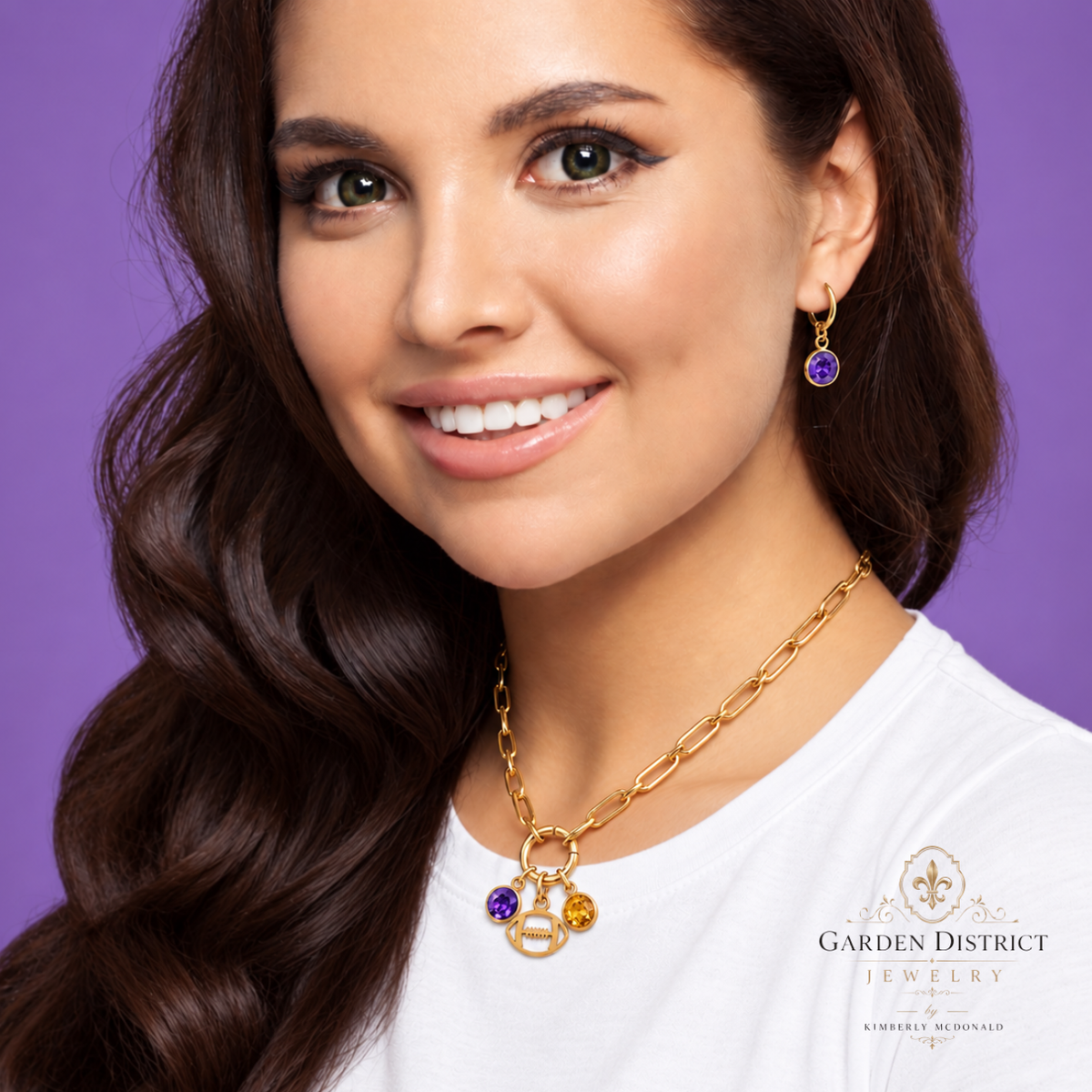 A woman with long dark brown hair models a gold chain necklace and matching gold drop earrings with purple and yellow gemstones against a solid purple background. The brand logo 'Garden District Jewelry' is visible on her white t-shirt.