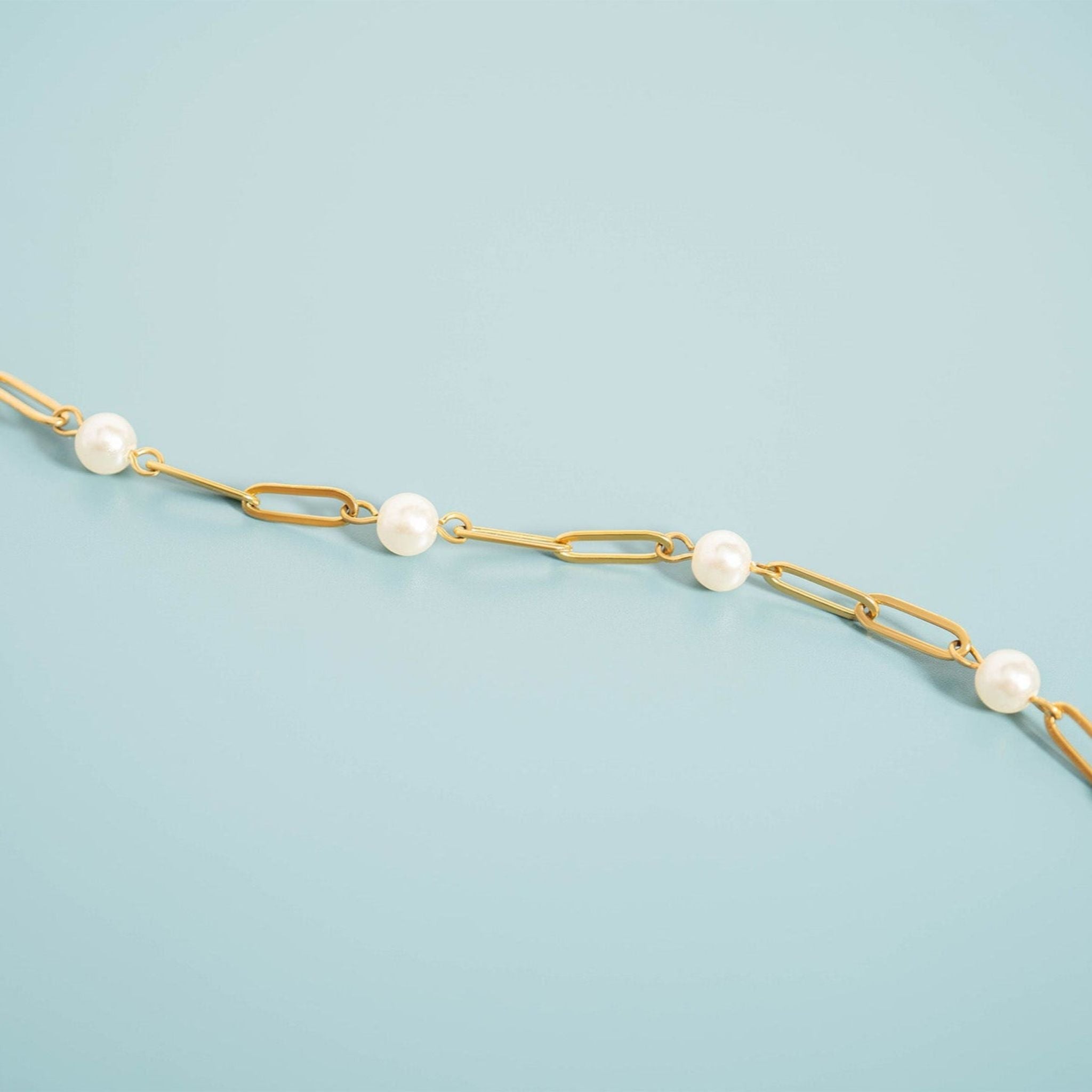 A gold paperclip chain necklace with evenly spaced white pearl beads, displayed diagonally on a light blue background.