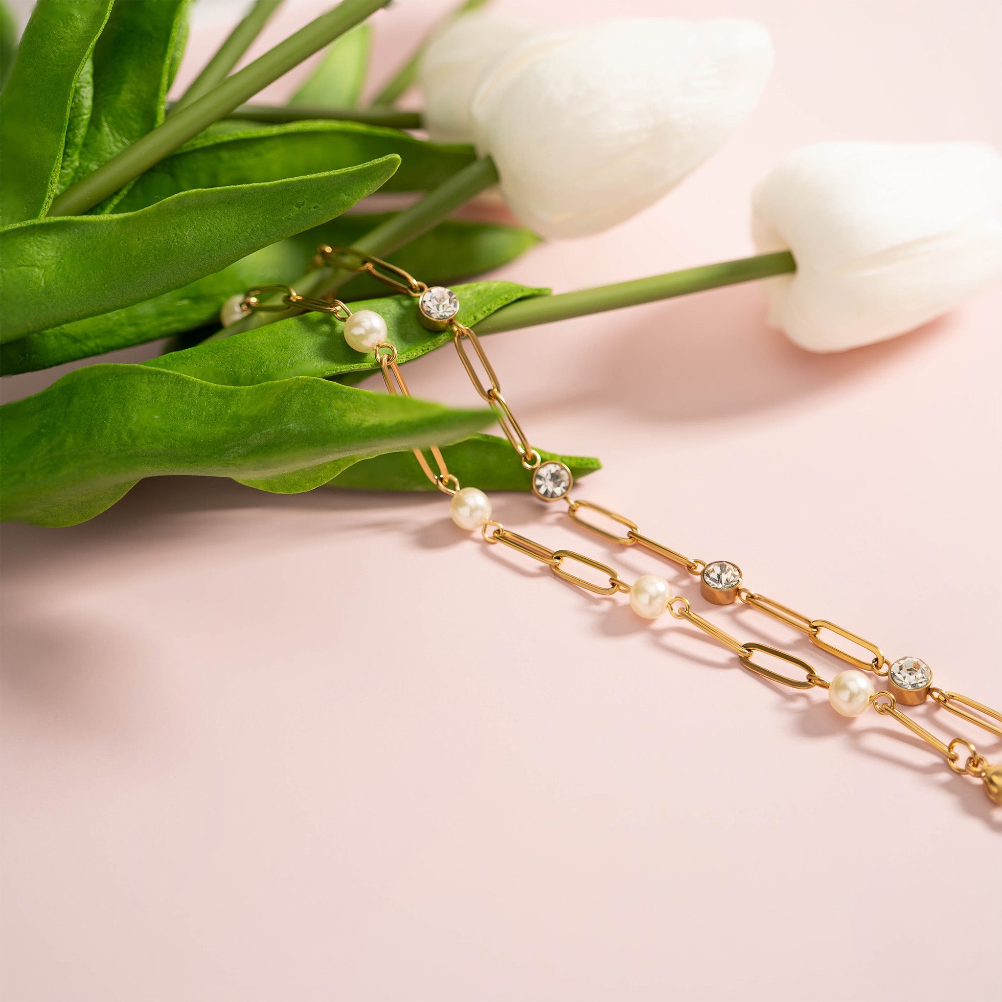 Gold chain necklace with pearls on a pink surface with green leaves and white flowers.