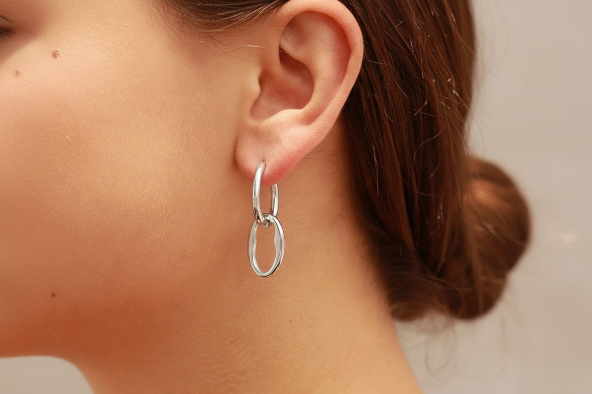 A close-up of a model's ear wearing silver hoop earring with a smaller interlocking oval drop, shown against skin and brown hair.