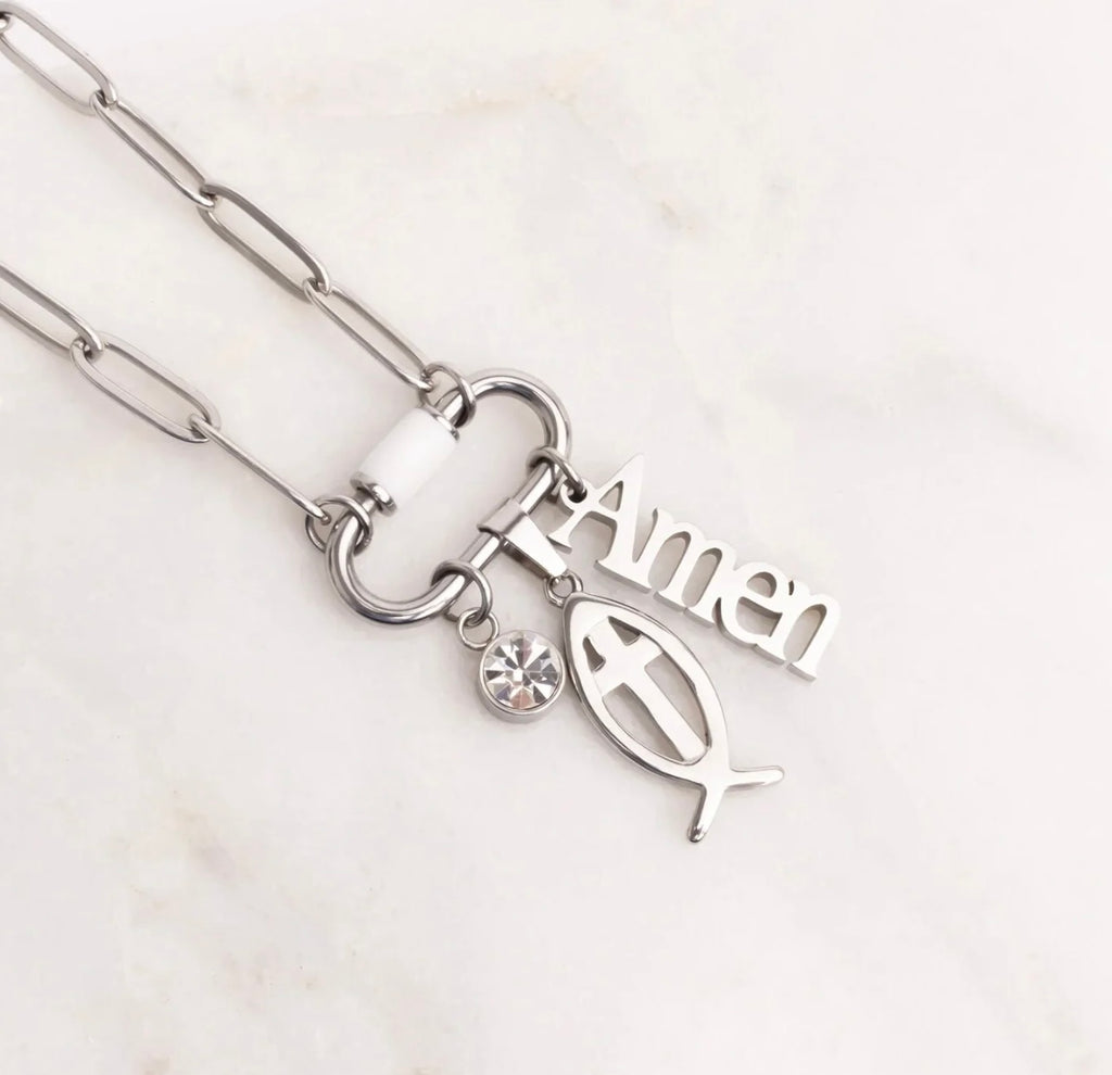 Silver paperclip chain necklace with a clasp featuring a white bead, a small clear rhinestone, an olive branch charm, and an 'Amen' word charm, displayed on a light marble background.
