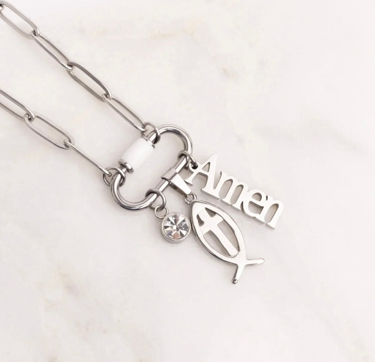 Silver paperclip chain necklace with a clasp featuring a white bead, a small clear rhinestone, an olive branch charm, and an 'Amen' word charm, displayed on a light marble background.