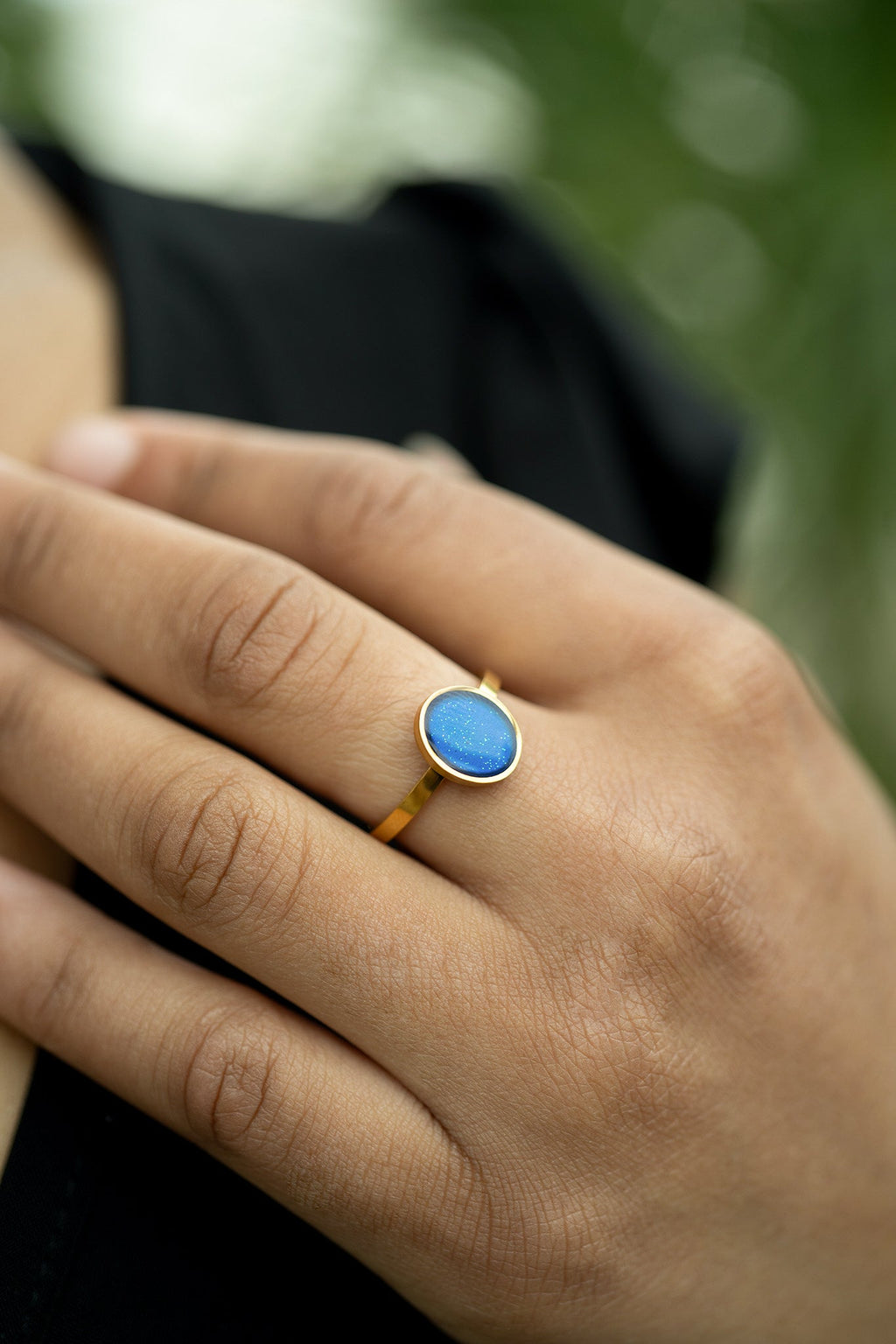 Hand wearing a gold ring with a blue stone against a blurred green background