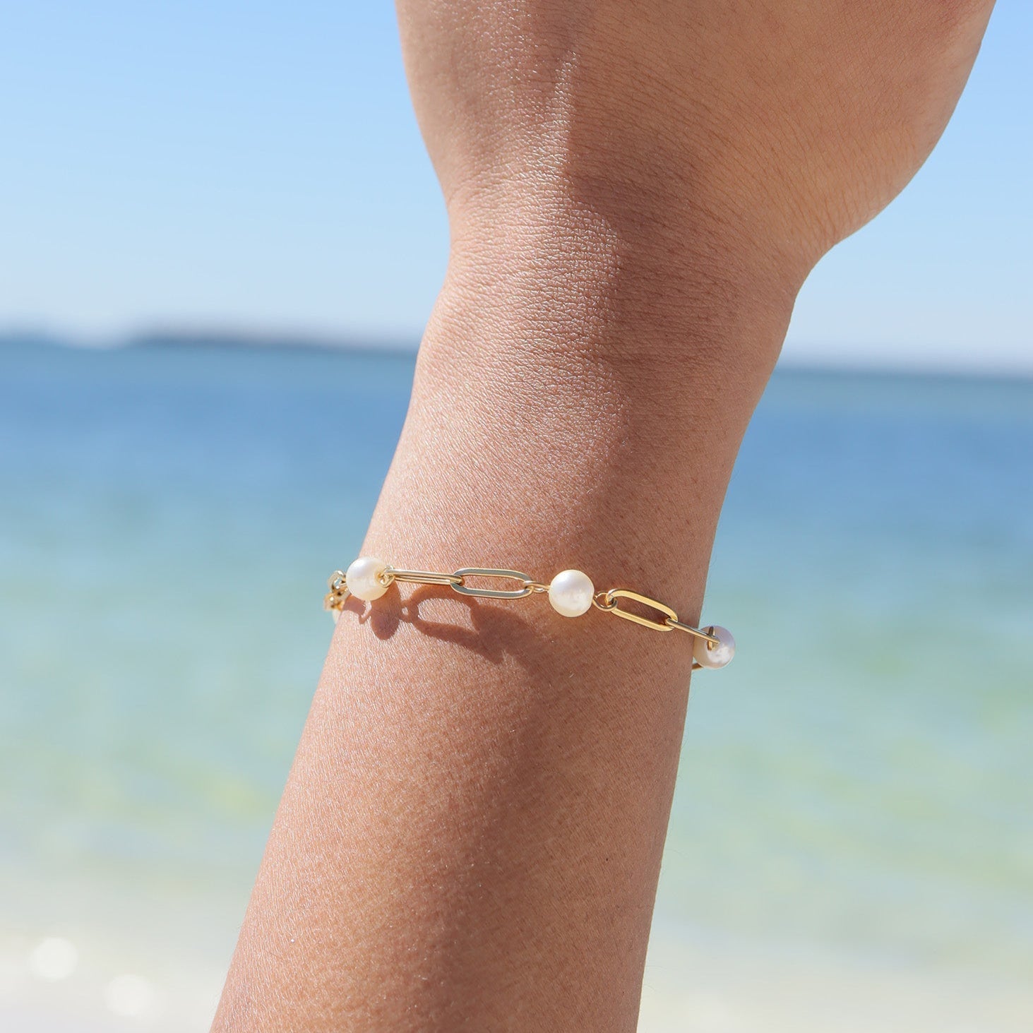 Gold bracelet with pearls on a person's wrist against a blurred beach background