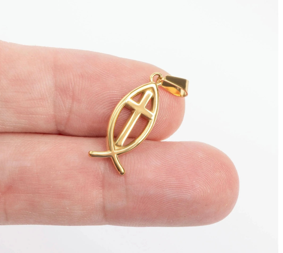 A small gold pendant shaped like a Christian fish symbol with a cross inside, shown resting on a person's fingers against a plain white background.