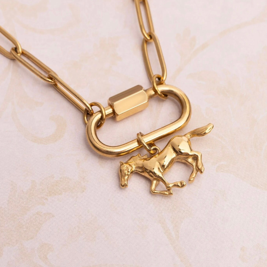 Gold-toned paperclip chain necklace with a toggle clasp and a small running horse charm, photographed on a pale pink patterned surface.