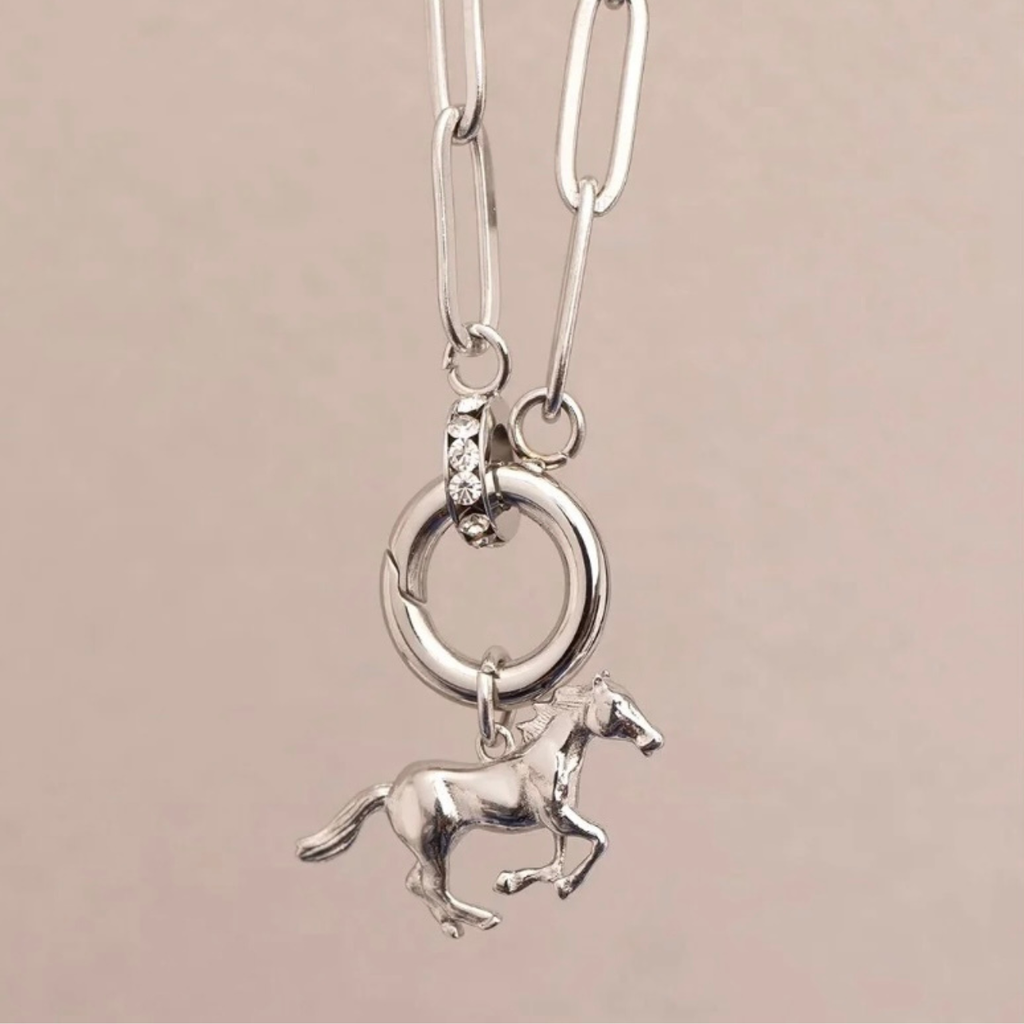 A silver paperclip necklace with a circular ring and a small running horse charm, shown against a solid light pink background.