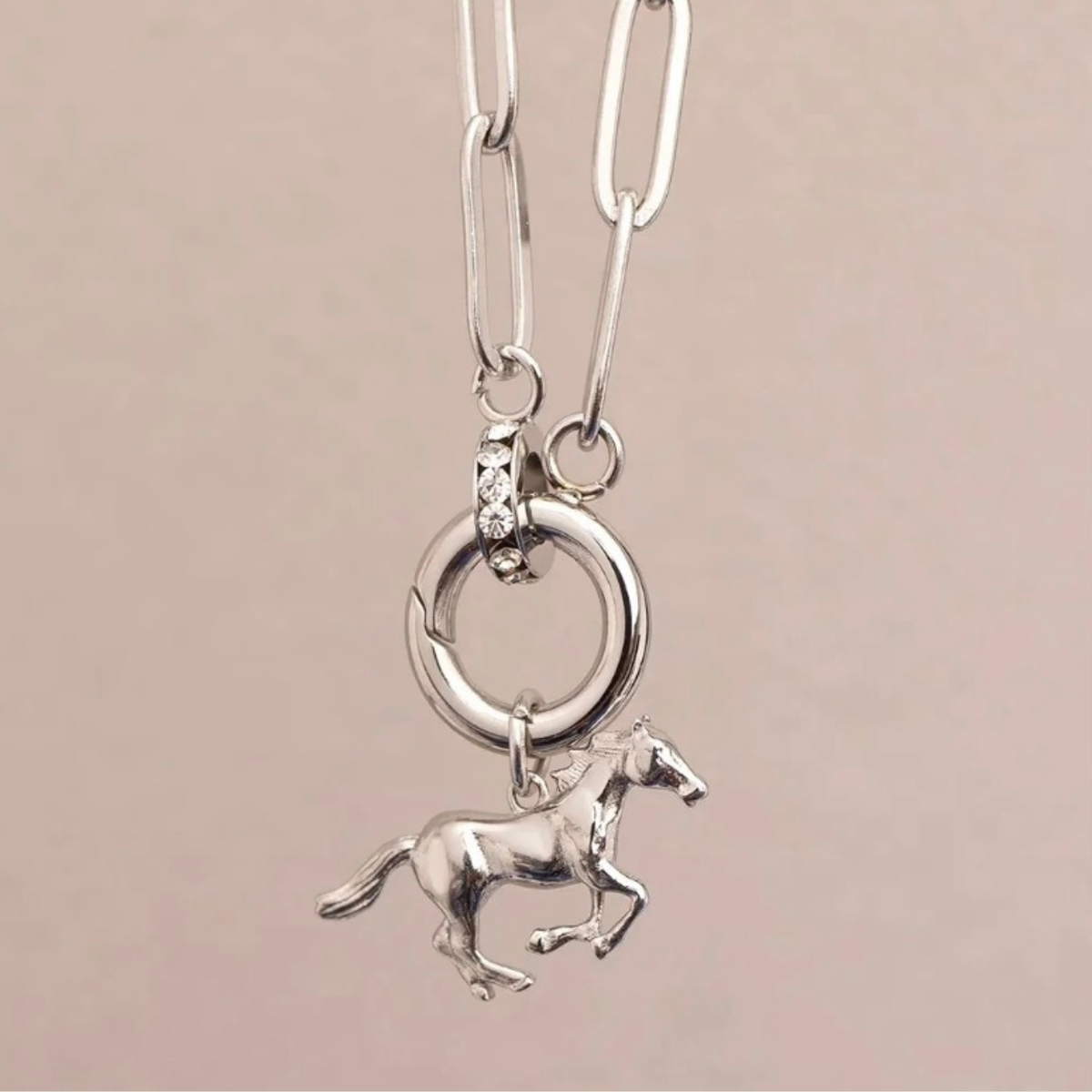 A silver paperclip necklace with a circular ring and a small running horse charm, shown against a solid light pink background.